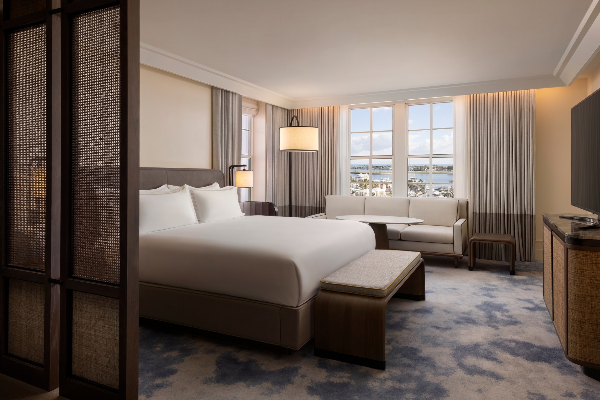 Spacious hotel guest room featuring a king-size bed with crisp white linens, a bench at the foot of the bed, a sofa seating area by multi-pane windows overlooking the water, and woven partition screens.
