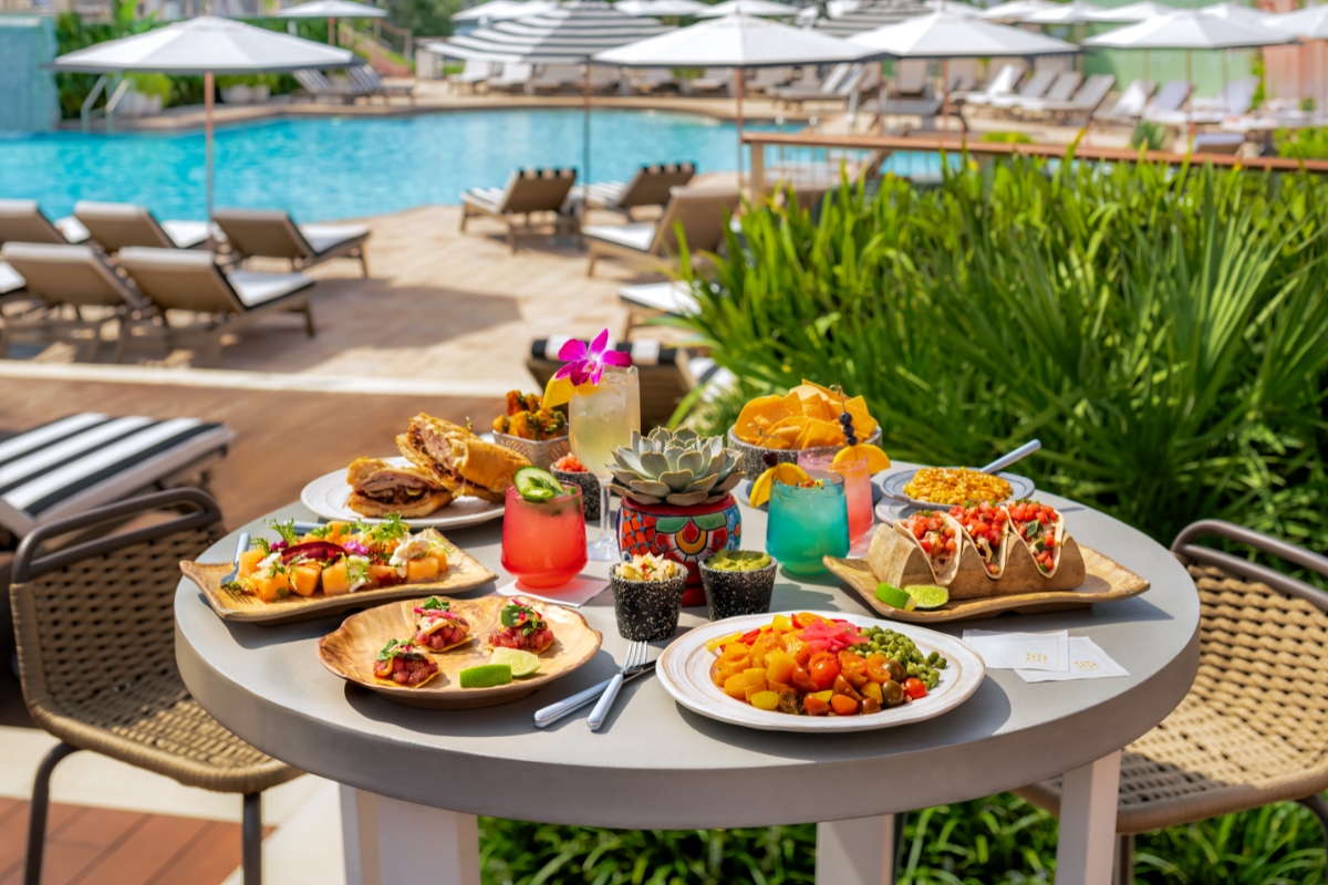 A round poolside patio table filled with a colorful array of food and cocktails, including tacos, sandwiches, appetizers, and vibrant drinks, with a resort pool and lounge chairs in the background.