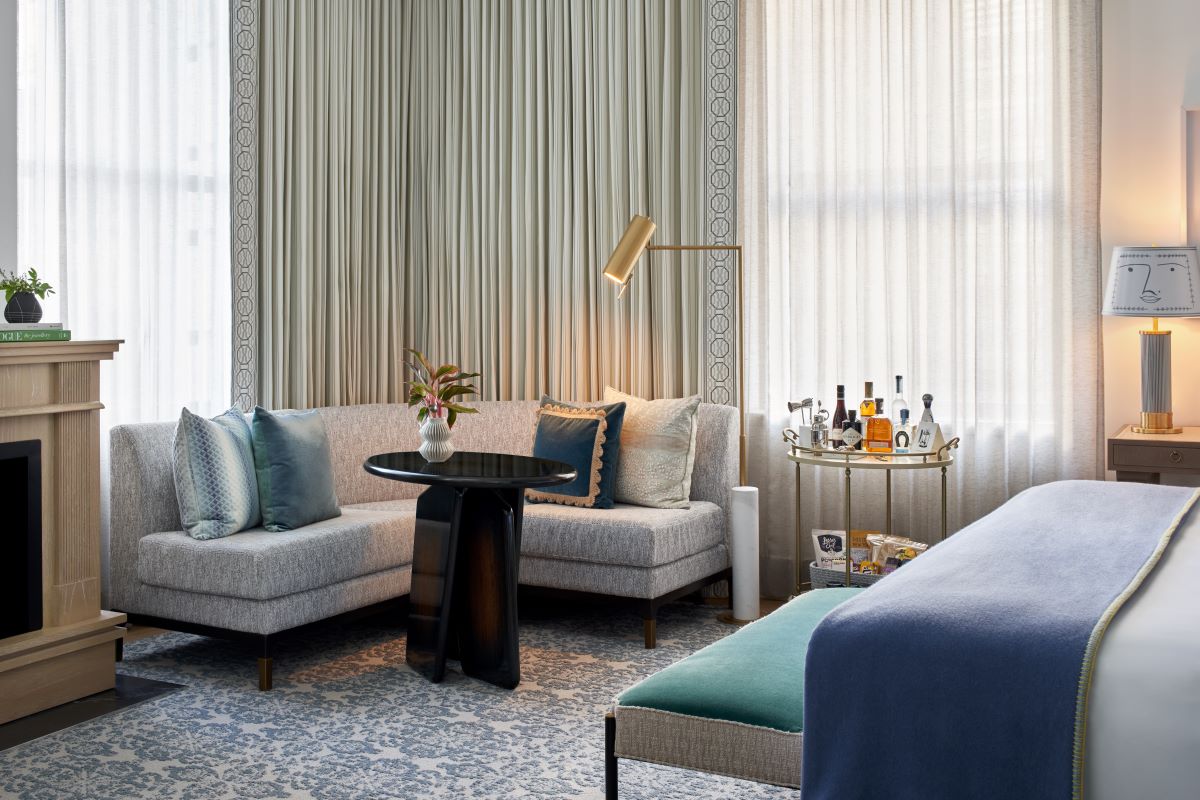 A cozy seating corner of a luxury hotel suite featuring a grey curved sectional sofa adorned with blue and patterned throw pillows, a dark round accent table, a brass floor lamp, and a fully stocked bar cart set against large sheer-curtained windows.