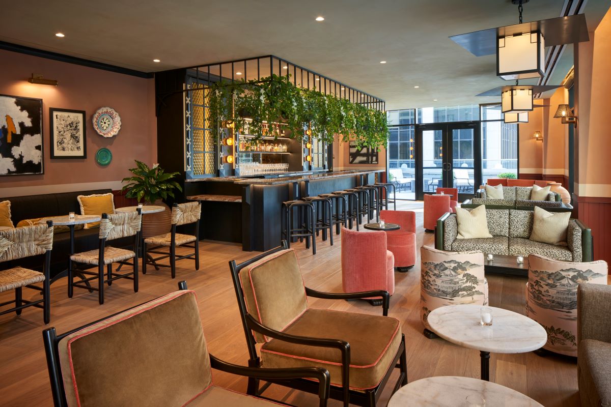 A stylish cocktail lounge and bar featuring mustard-yellow velvet armchairs, coral barrel armchairs, patterned couches, and woven dining chairs arranged on light wood flooring around marble coffee tables, with a dark steel bar counter framed by hanging gr