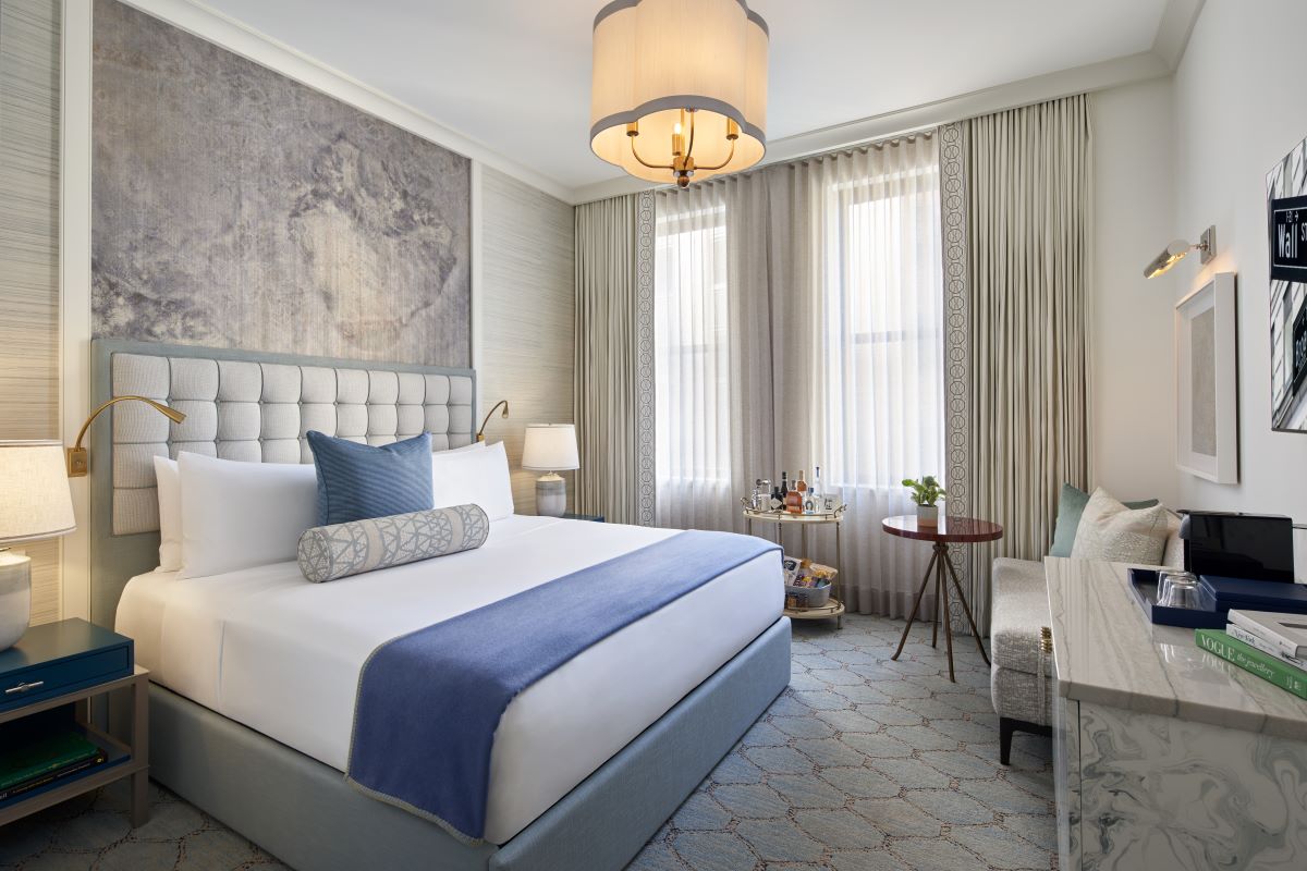 A bright hotel room featuring a king bed with a tufted headboard, blue bed throw, and accent pillows, set against a grey textured wall feature, flanked by blue nightstands, bright windows with sheer drapes, and a small seating nook with a round table and 