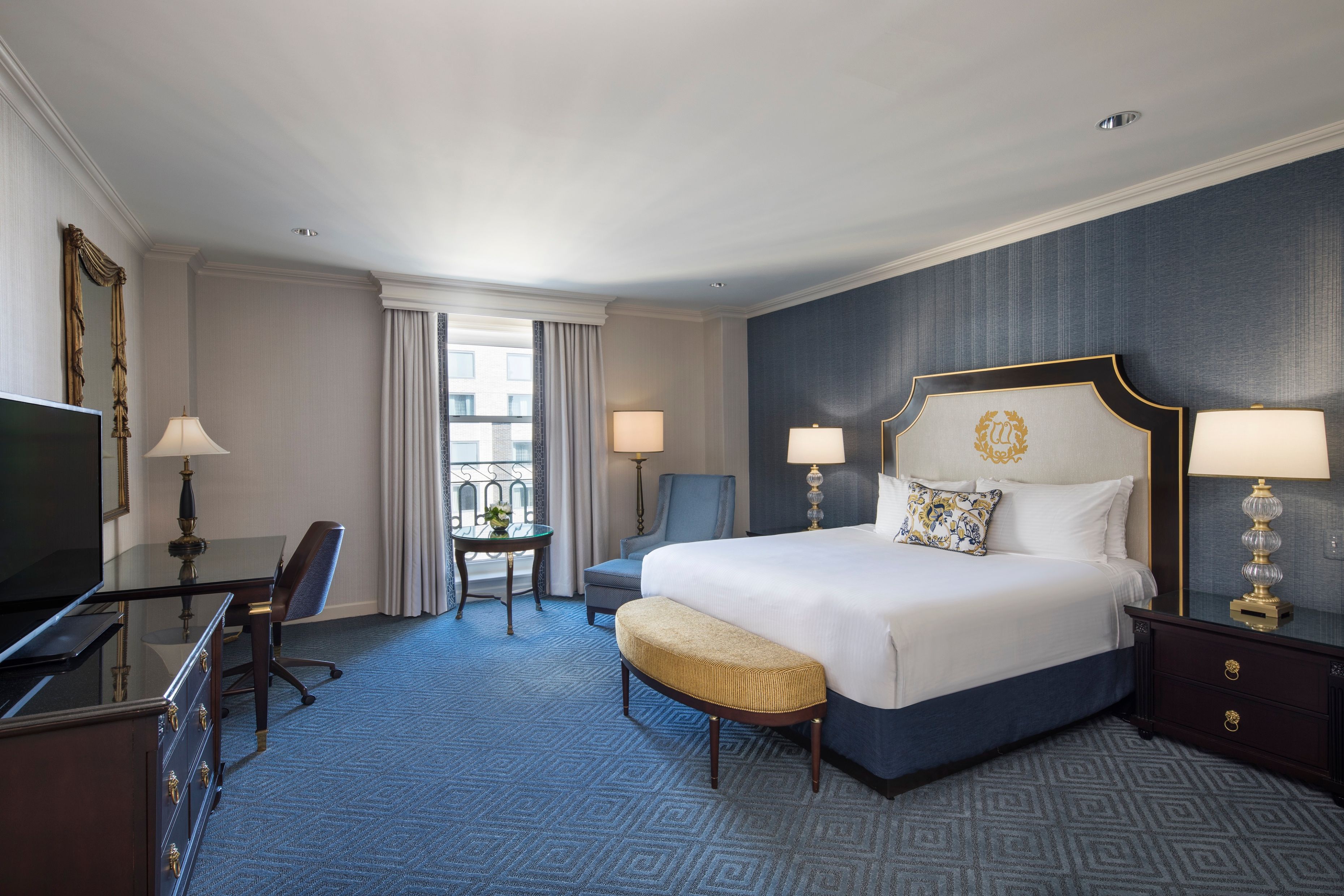 Spacious hotel guest bedroom featuring a king bed with an upholstered headboard, blue accent wall, blue patterned carpet, dark wood furniture including a desk and dresser, and a window with light drapes.