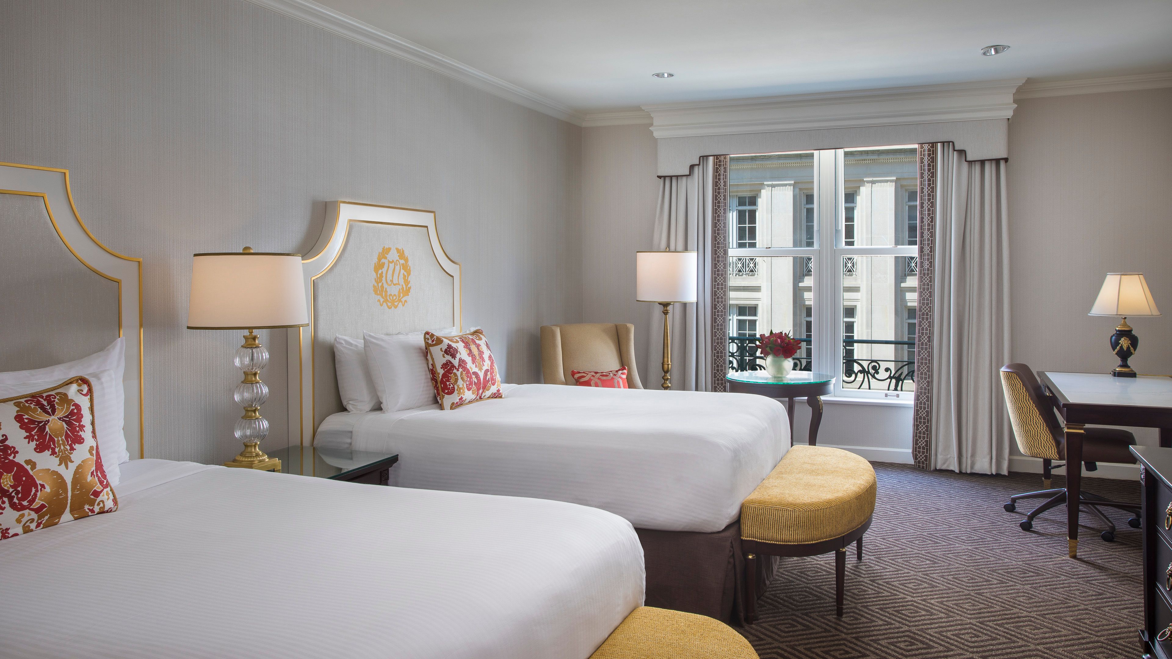 Hotel guest room with two beds featuring gold-trimmed white upholstered headboards, red patterned throw pillows, yellow padded benches at the foot of each bed, and a large window looking out onto historic architecture.