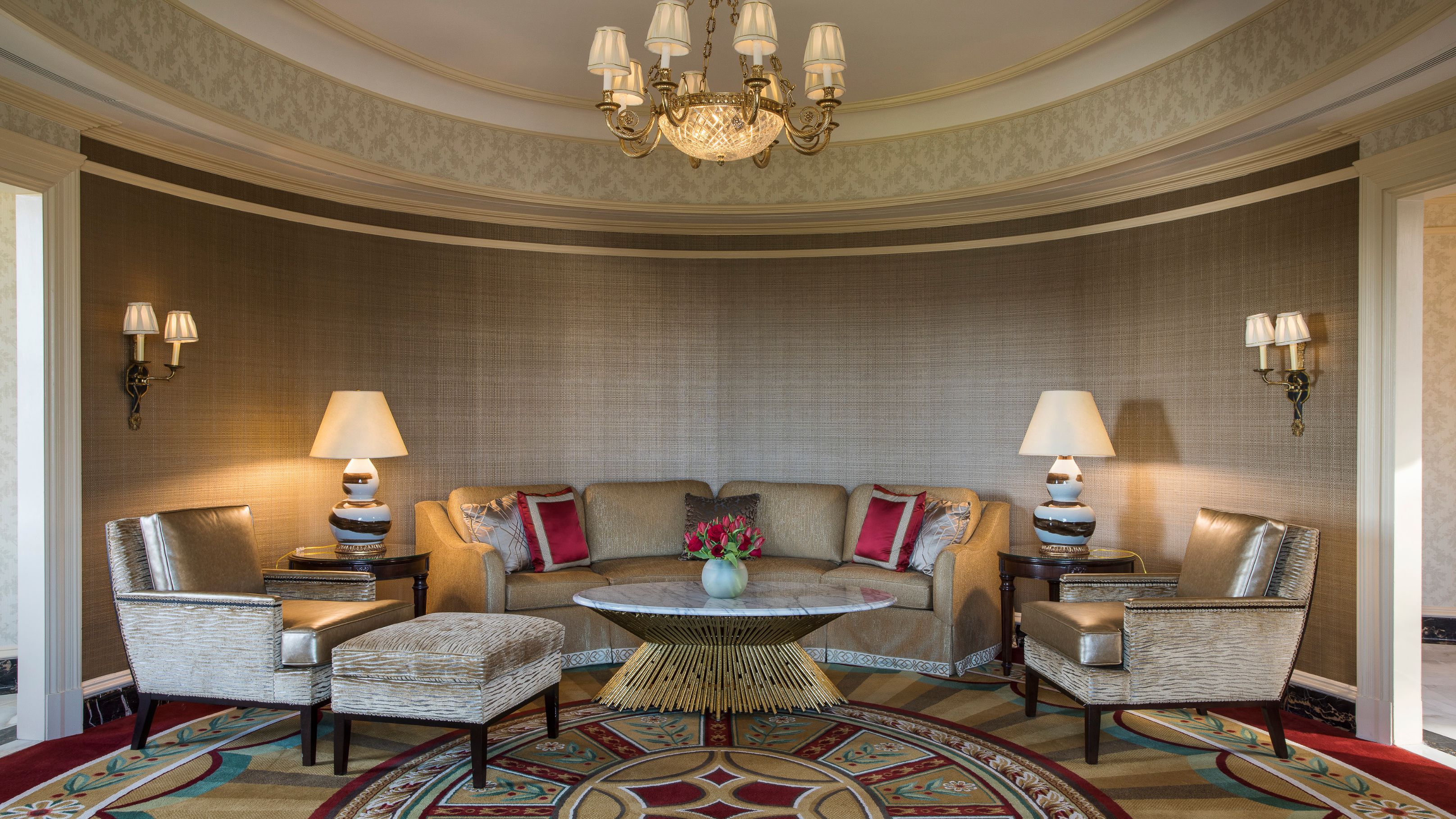 An elegant, curved rotunda sitting area with textured warm gray grasscloth wallpaper, a beige curved sectional sofa, metallic armchairs, a central round marble coffee table with a gold base, and soft lighting from sconces and lamps.