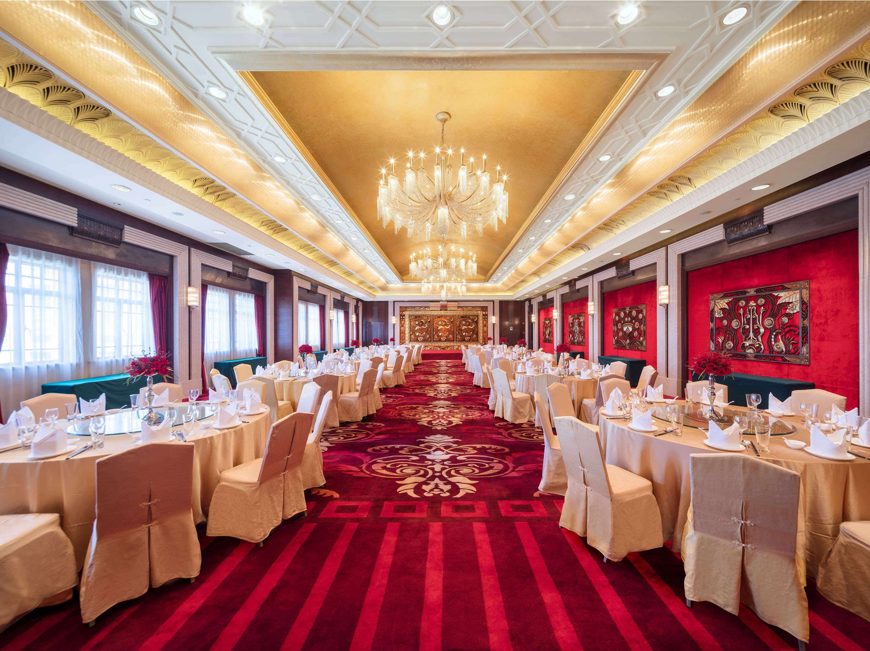 A grand, opulent ballroom featuring a rich red patterned carpet, rows of round banquet tables with gold chair covers and formal place settings, glowing crystal chandeliers, and a gold-leaf coffered ceiling.