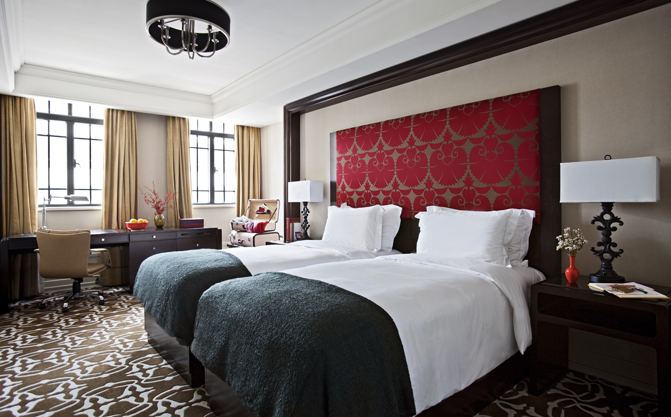 A stylish hotel room featuring two twin beds with white linens and dark green throws beneath a tall red patterned upholstered headboard, a work desk by multi-pane windows with beige drapes, and an ornate patterned carpet.