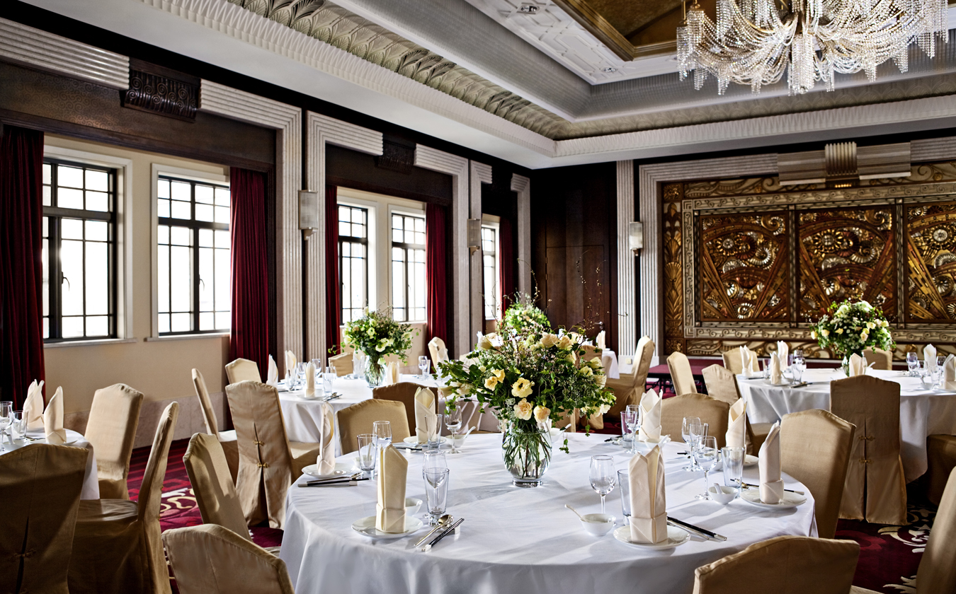 A lavish banquet room set up with round dining tables draped in white tablecloths, gold-sheathed chairs, elegant floral centerpieces, crystal chandeliers, and opulent gold Art Deco wall paneling.