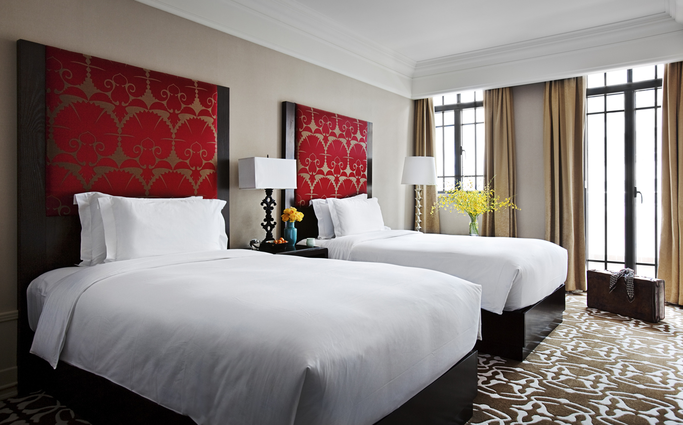A elegant hotel room featuring two beds with crisp white duvets, each with a tall dark wooden frame and a red patterned fabric headboard insert, set on a patterned carpet near French doors with beige drapes.