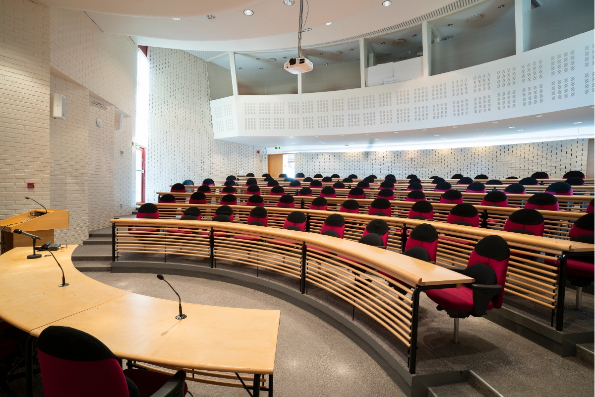 A large, modern amphitheater-style conference auditorium featuring tiered rows of light wood desks with slats, red and black office chairs, white textured brick walls, a curved speaker's podium table with microphones in the foreground, and a projector