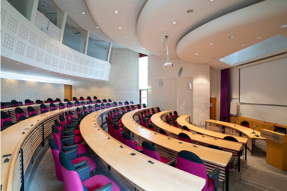 A wide-angle view of a modern tiered auditorium with curved light wood desk rows, pink and black upholstered seating, white brick walls, a wooden podium, a large projection screen at the front, and floating ceiling panels with recessed lighting.