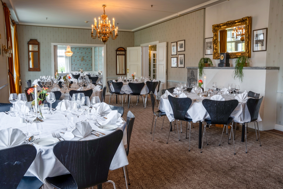 An elegant dining room set for a banquet or event, featuring round tables with white tablecloths, fine stemware, folded white napkins, black Arne Jacobsen Series 7-style chairs, classic crystal chandeliers, and a gold ornate mirror above a mantlepiece.