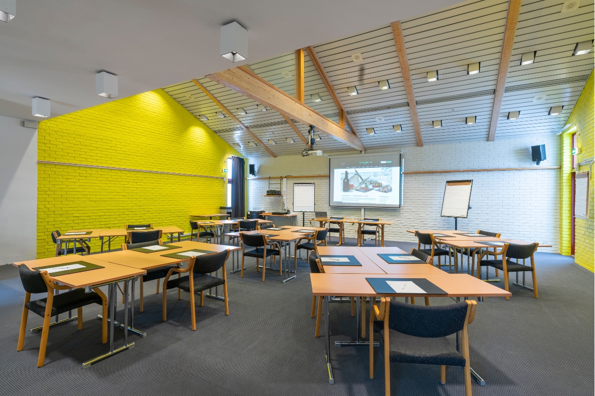A bright conference room set up with small wooden tables and dark chairs, featuring a bold vibrant yellow accent brick wall, high vaulted ceiling with exposed wooden beams, white painted brick walls, flipcharts, and a projector screen at the front.