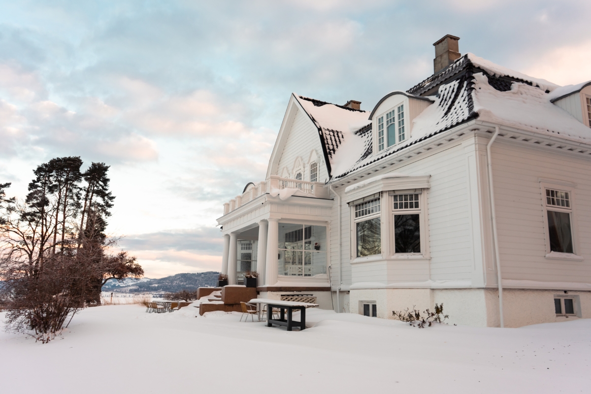 A grand white two-story historic manor covered in snow during winter, featuring classical columns, dormer windows, a snow-laden roof, and a scenic landscape view under a soft pastel sky.