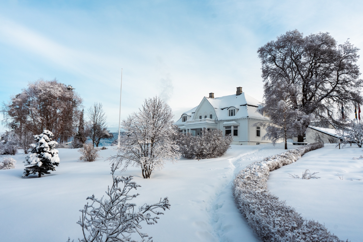 A wide snow-covered estate featuring a white wooden manor house nestled among frost-covered trees, with a winding snowy path bordered by low snow-draped hedges under a pale blue winter sky.