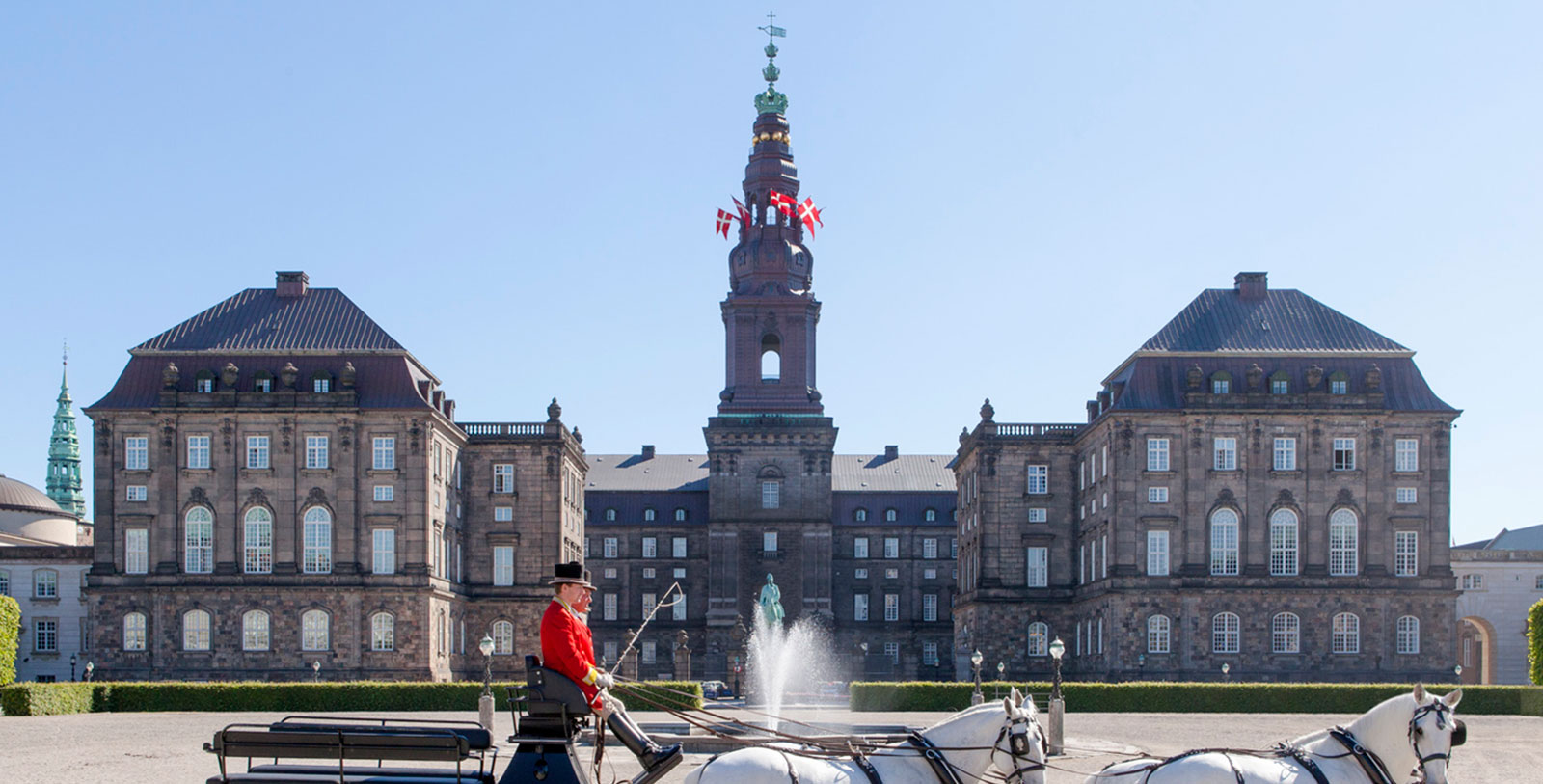 a man in a black hat sitting on a horse carriage in front of a large building with Christiansborg Palace in the background