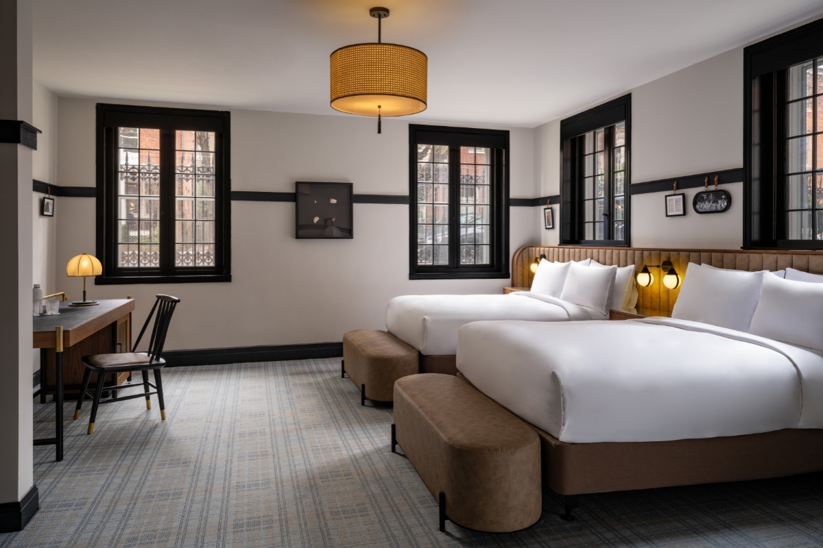 A stylish hotel bedroom with two queen beds featuring white linens, light brown upholstered bases and matching footrest benches, black-framed windows, a writing desk with a lamp, plaid carpeting, and a woven drum pendant light hanging from the ceiling.