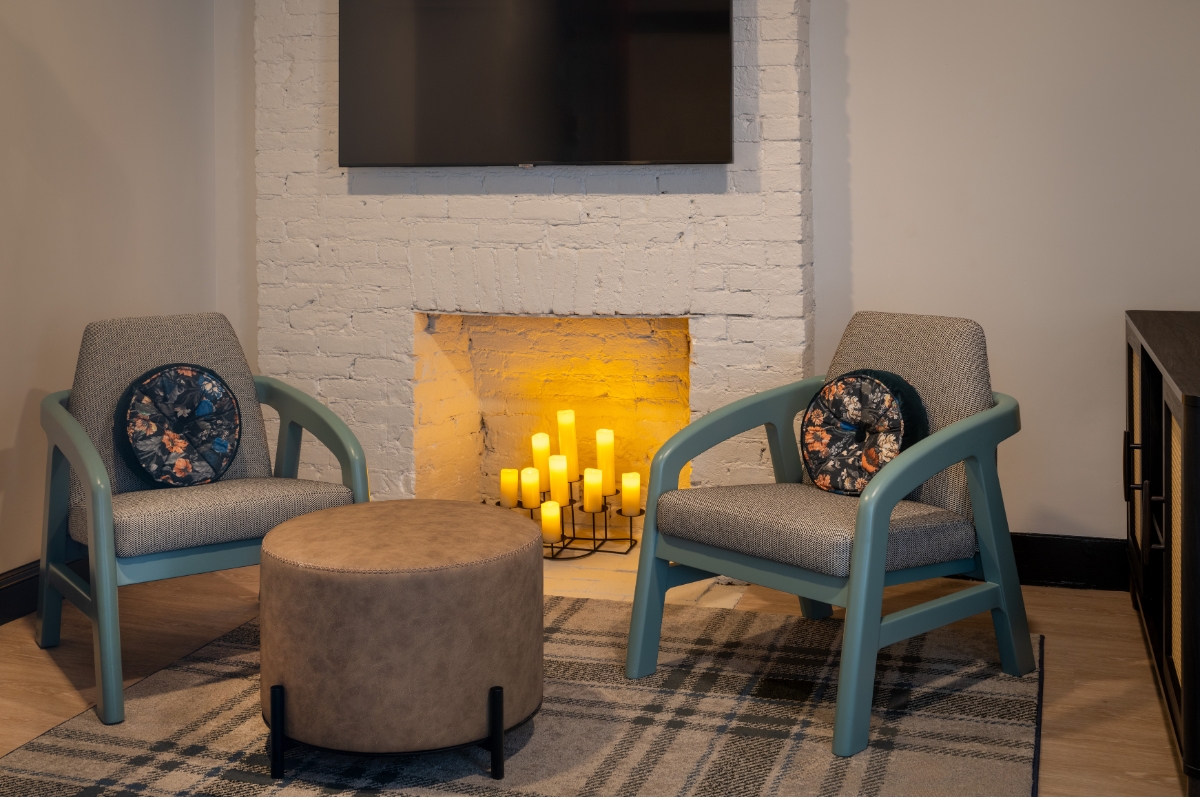 A cozy lounge area featuring two grey upholstered armchairs with teal wooden frames, round floral accent pillows, a round leather ottoman, and a plaid rug in front of a white painted brick fireplace glowing with pillar candles.