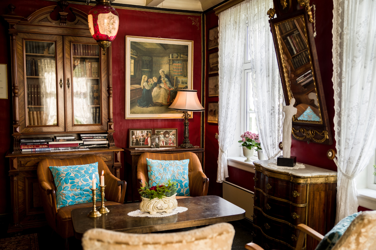 An richly decorated vintage room featuring deep red walls, a large wooden hutch filled with books, two armchairs with bright blue patterned pillows facing a coffee table with candles, sheer lace curtains, and classic artwork.