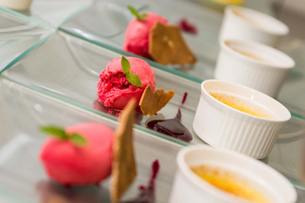 A row of plated desserts on clear glass plates, each featuring a scoop of vibrant pink sorbet garnished with a mint sprig, a crisp wafer, a drizzle of berry sauce, and an accompanying white ramekin filled with creme brulee.