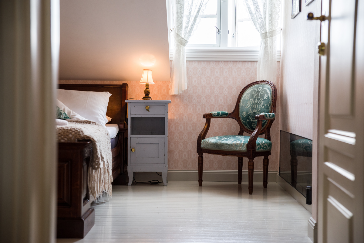 A cozy, vintage-style bedroom with light pink patterned wallpaper, a dark wooden bed with white linens and a throw blanket, a small grey wooden nightstand topped with a glowing lamp, and a green patterned armchair under a window with sheer white curtains.