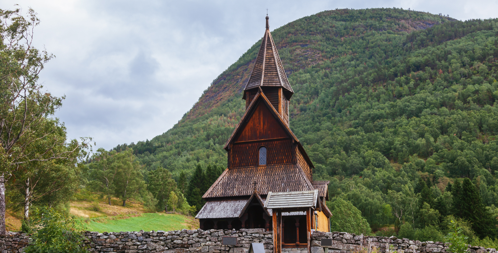 a wooden church with a stone wall and a hill in the background with Urnes Stave Church in the background