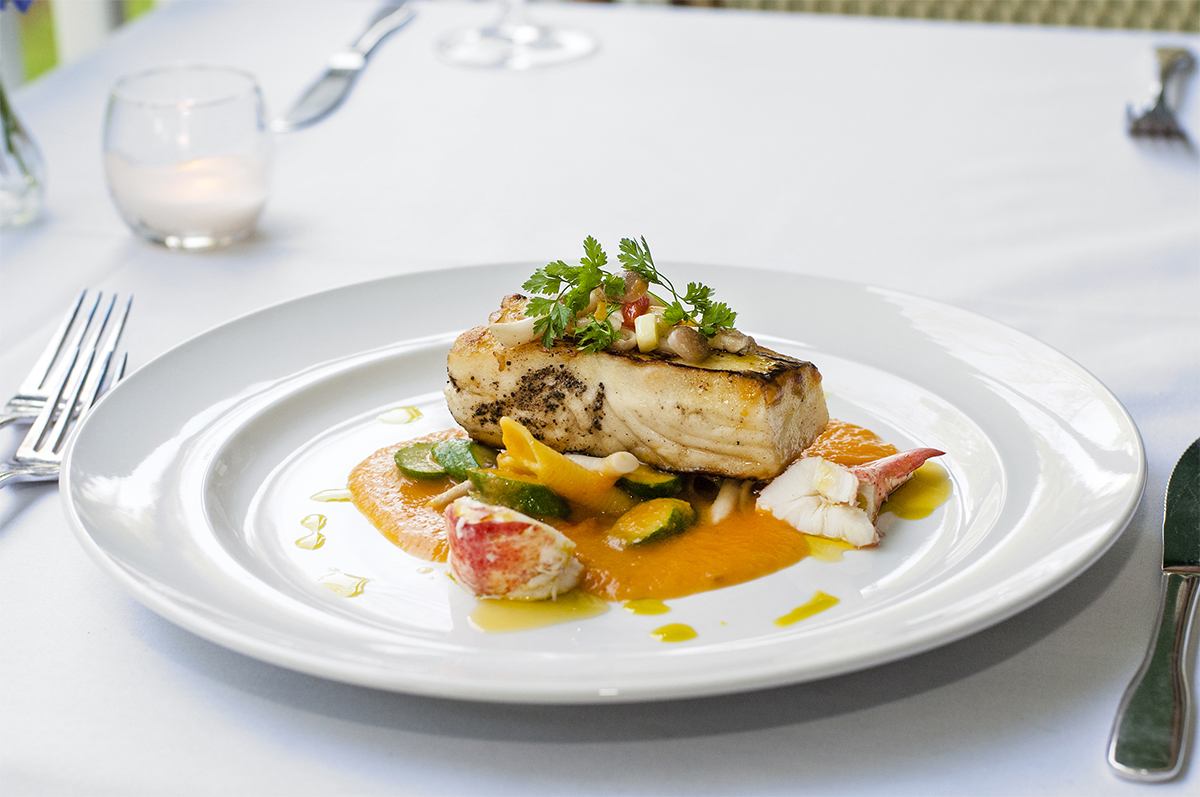 Gourmet plated seafood dish featuring a seared white fish fillet topped with herbs, resting over lobster meat, sliced squash, and an orange puree sauce on a white dinner plate.