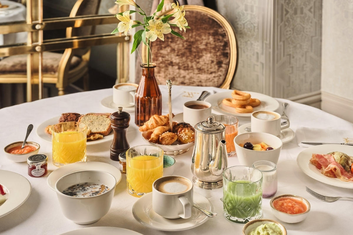 An elegant dining table set with a lavish breakfast spread, including fresh pastries, churros, coffee cups, fruit juices, yogurt, silver coffee pots, and a small flower vase.
