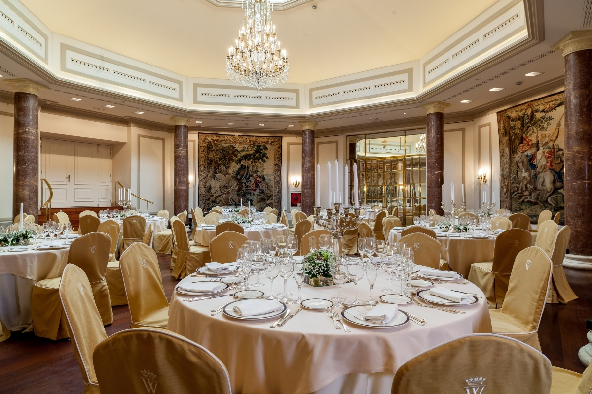 A grand octagonal banquet hall prepared for a formal event, with round tables draped in cream linens, gold chair covers, tall candelabras, classic marble columns, historic wall tapestries, and a central crystal chandelier.