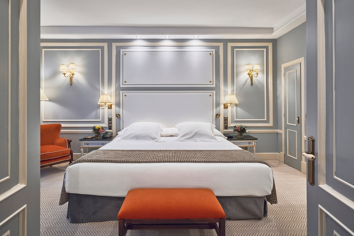 A refined hotel bedroom framed through open doors, showcasing light grey walls with classic wainscoting, a king bed accented by an orange bench, and matching orange armchair.
