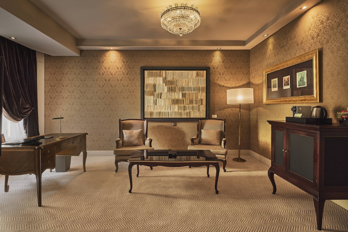 A luxury executive hotel suite featuring warm damask wallpaper, a crystal chandelier, two upholstered armchair seats with a coffee table, a classic wooden writing desk, and a side cabinet.