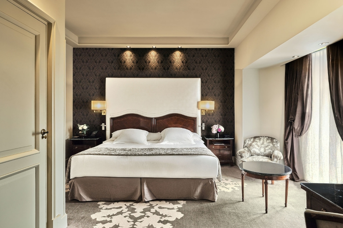 A sophisticated luxury bedroom featuring a plush bed with an oversized white headboard set against a dark damask accent wall, flanked by warm sconces and dark velvet curtains.