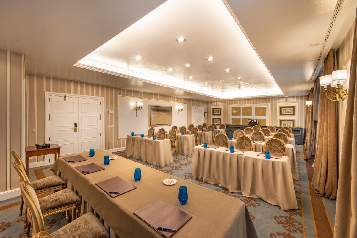 An elegant conference room set up in classroom style with beige-draped tables, gold oval-backed upholstered chairs, blue drinking glasses, striped walls, and a recessed ceiling panel with warm lighting.